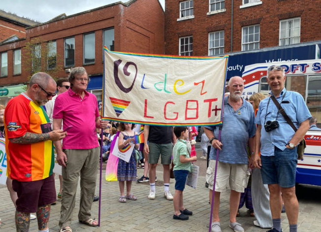 A group of four individuals holding a banner that reads 'Older LGBT+' at a Pride event, surrounded by a colorful crowd.