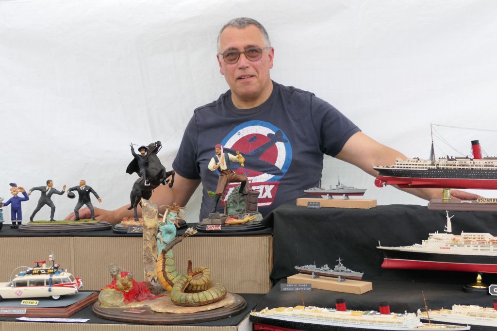 A man sitting behind a display of various model figures and ships, showcasing detailed miniatures at an event.