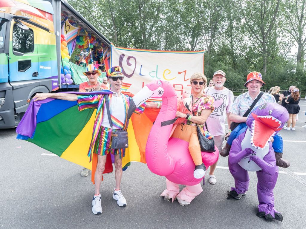 A group of six people celebrating at a Pride event, wearing colorful outfits and accessories. One person holds a large rainbow flag, while others are dressed in inflatable costume animals. They pose in front of a brightly decorated truck with balloons and a banner.