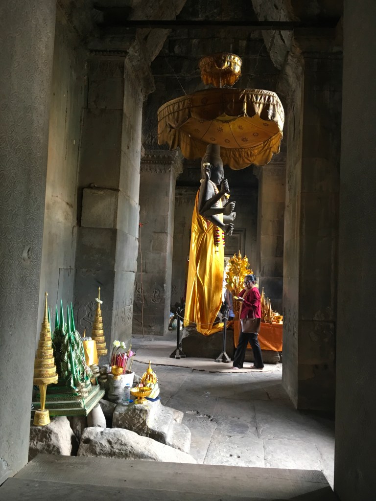 A view of a temple interior featuring a large black statue of a deity draped in gold fabric. Several offerings and decorations are placed in front of the statue. A person is walking through the doorway, observing the scene.