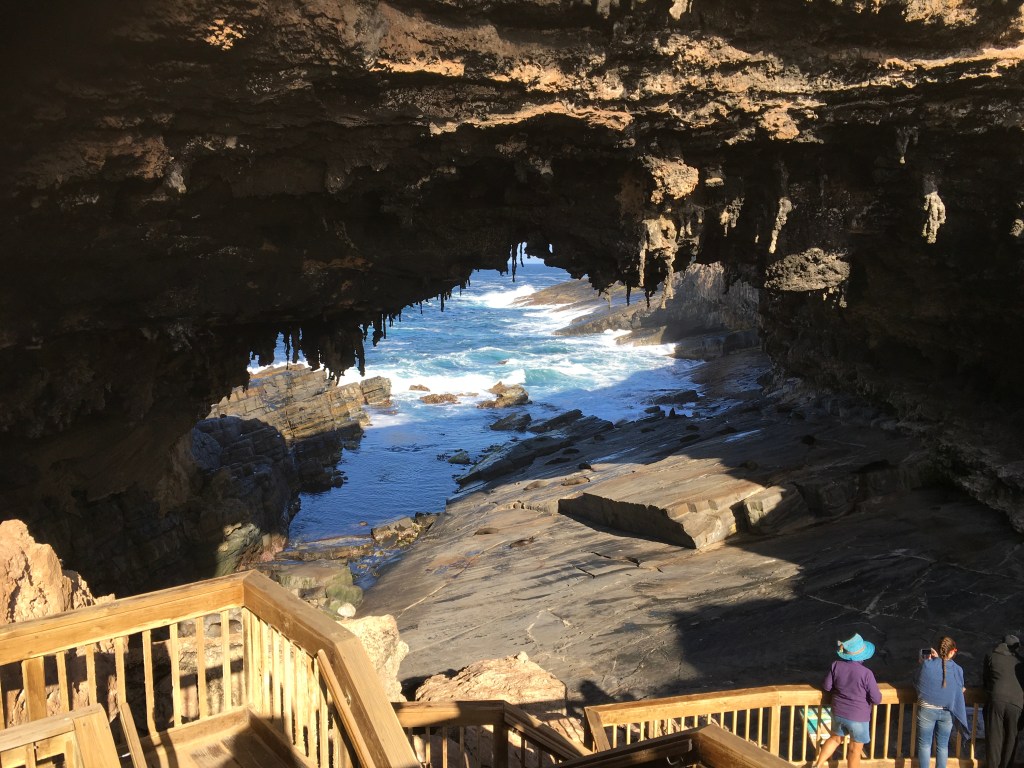 View from a cave opening overlooking rocky coastline and ocean waves, with visitors observing from a wooden staircase.