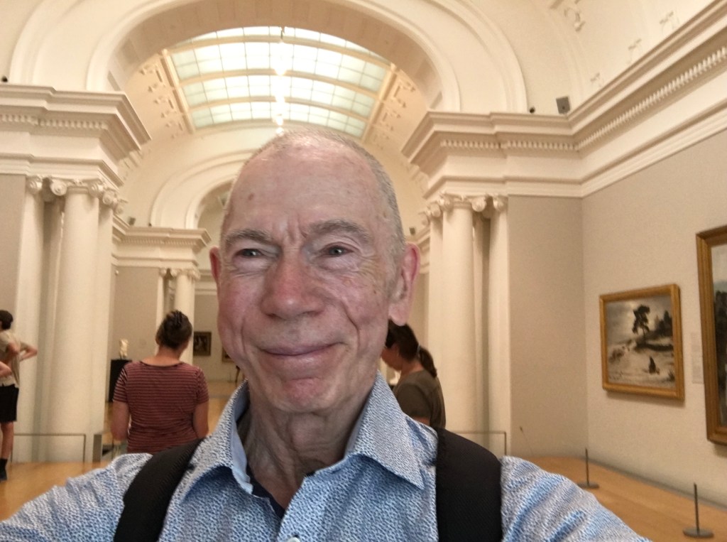 A man smiling for a selfie in an art museum, with sculptures and paintings visible in the background.