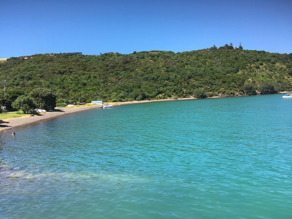 A scenic view of a calm turquoise bay with a beach. Lush green hills line the background. A few small boats are visible on the water, and a person is standing by the shore.