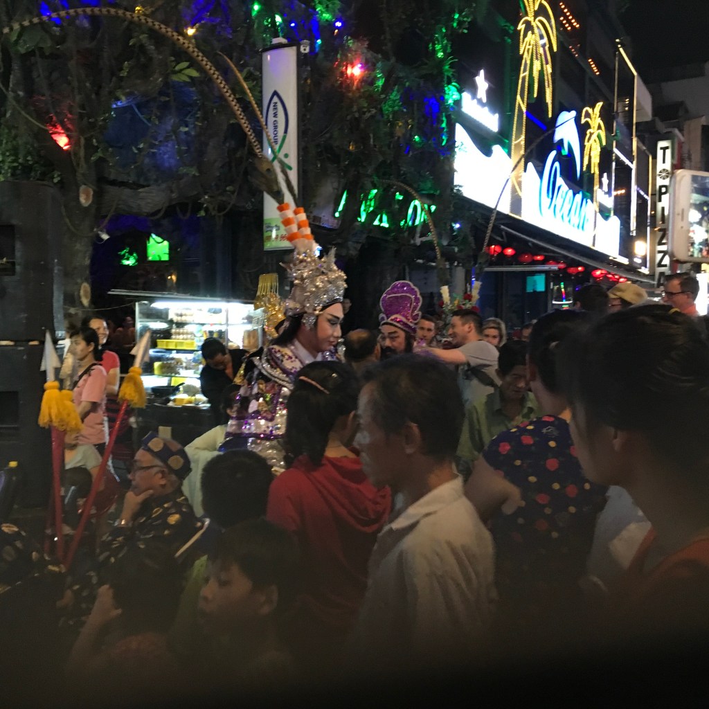 A lively street scene at night featuring performers in traditional costumes surrounded by a crowd, colorful lights in the background, and street vendors.