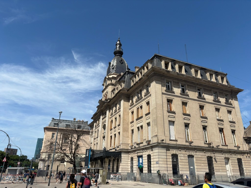 Historic building with ornate architecture and a tall tower, set against a clear blue sky, with pedestrians and street activity in the foreground.