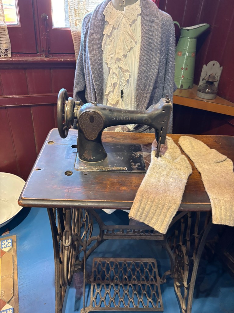 An antique Singer sewing machine on a wooden table with a pair of knitted gloves beside it, in front of a mannequin dressed in a vintage blouse and cardigan.