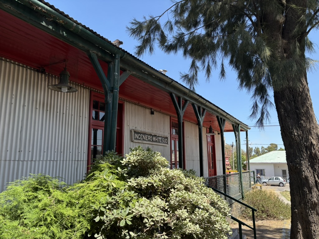 Exterior view of a building with a corrugated metal facade, featuring red trim, large windows, and a sign that reads 'Incenero White Sud,' surrounded by greenery and trees.