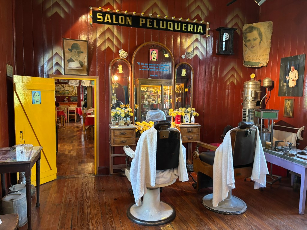 Interior of a vintage barbershop named 'Salon Peluqueria,' featuring two barber chairs with white capes, yellow door, and decorations including flowers and old photographs on the walls.