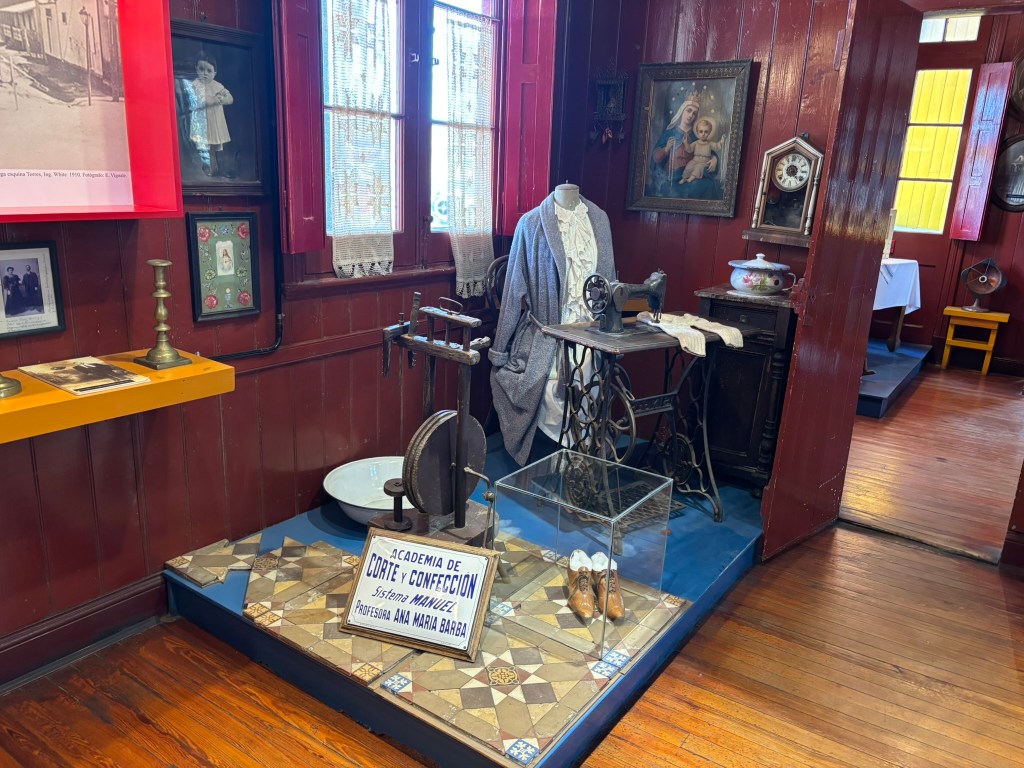 A vintage sewing machine and a dress on display in a room decorated with historical photographs and artifacts, along with a sign for a sewing academy.