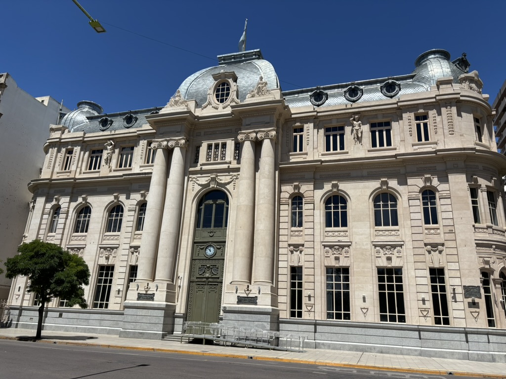 A grand historic building featuring classical architecture, with large columns, ornate sculptures, and a dome roof. The entrance has a clock and intricately detailed facade, surrounded by bright blue skies.