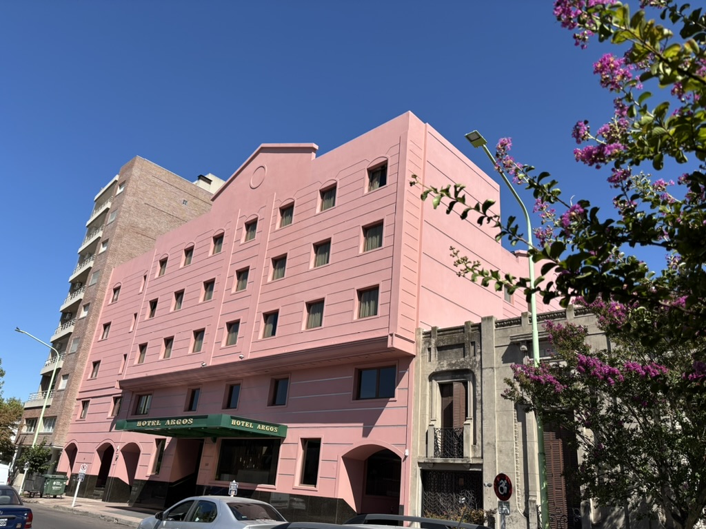 A large pink hotel building with several windows, featuring the name 'Hotel Argos' prominently displayed above the entrance. Nearby, there are smaller buildings and greenery in a clear blue sky.