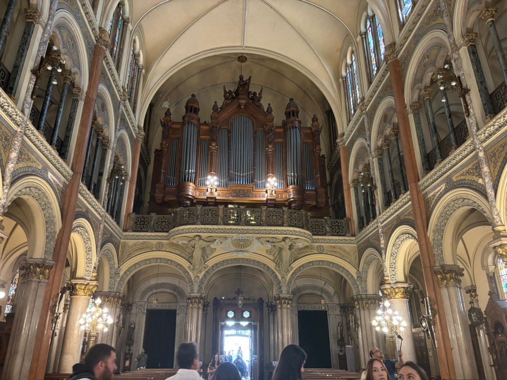 Interior view of a grand church featuring a large ornate organ at the front, surrounded by decorative arches and stained glass windows, with visitors admiring the architecture.