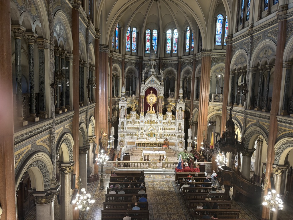 Interior view of a grand church, featuring intricate architecture, stained glass windows, and a central altar with ornate decorations. Several pews are filled with worshippers.