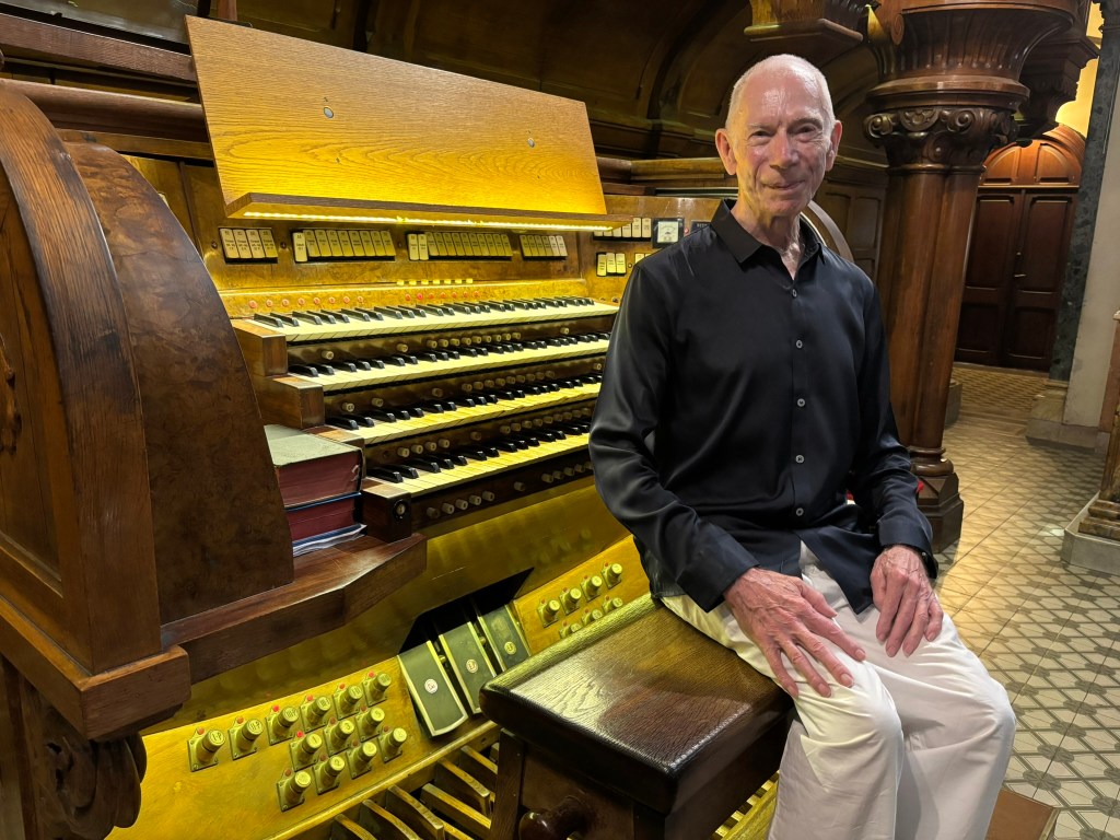 A seated man in a black shirt and white pants smiles beside a large ornate church organ, showcasing its keys and controls in a beautifully lit interior.