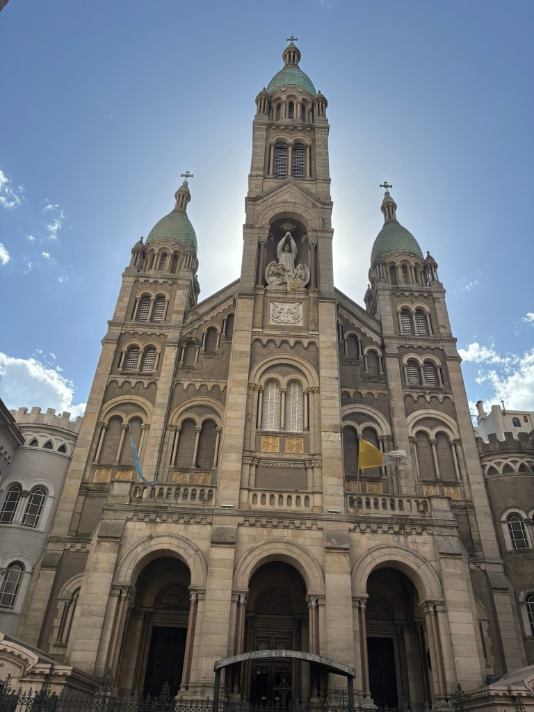 Facade of a large cathedral with three towers, featuring ornate stone details and a statue of a religious figure, against a blue sky with scattered clouds.