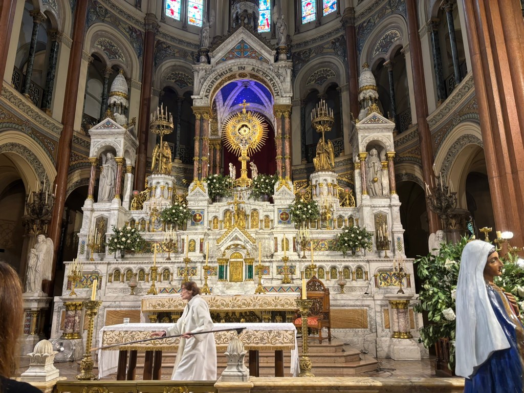 A beautifully decorated church altar featuring ornate marble work, gold accents, and floral arrangements. In the foreground, a person in a white robe is performing a religious ritual, with a statue of a saint visible on the right.