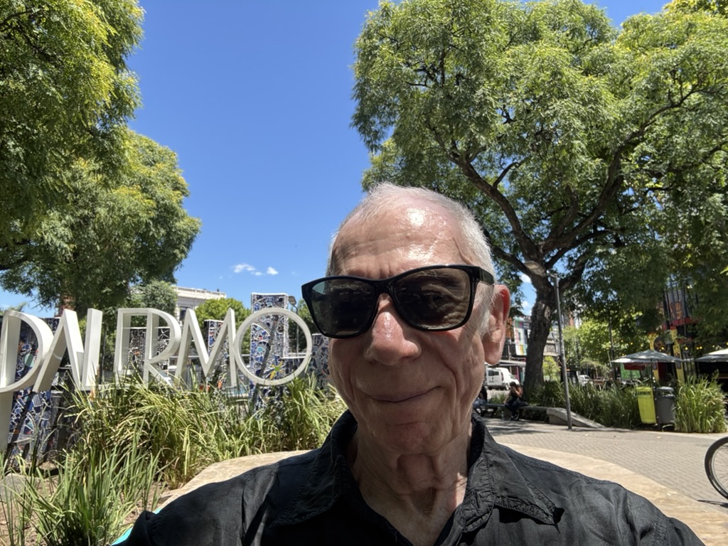 A person wearing sunglasses smiles in front of a sign reading 'Palermo' surrounded by greenery and trees on a sunny day.
