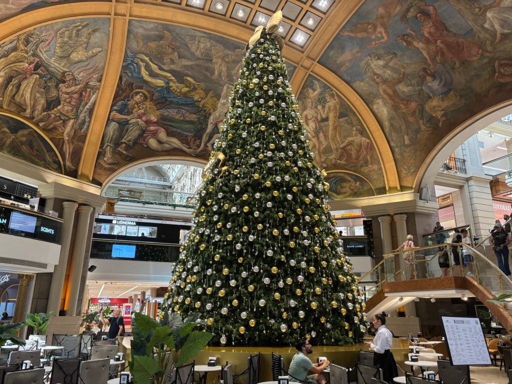 A large Christmas tree decorated with gold and silver ornaments stands in a shopping center, beneath a beautifully painted ceiling. People are seen in the space, with tables and chairs arranged nearby.