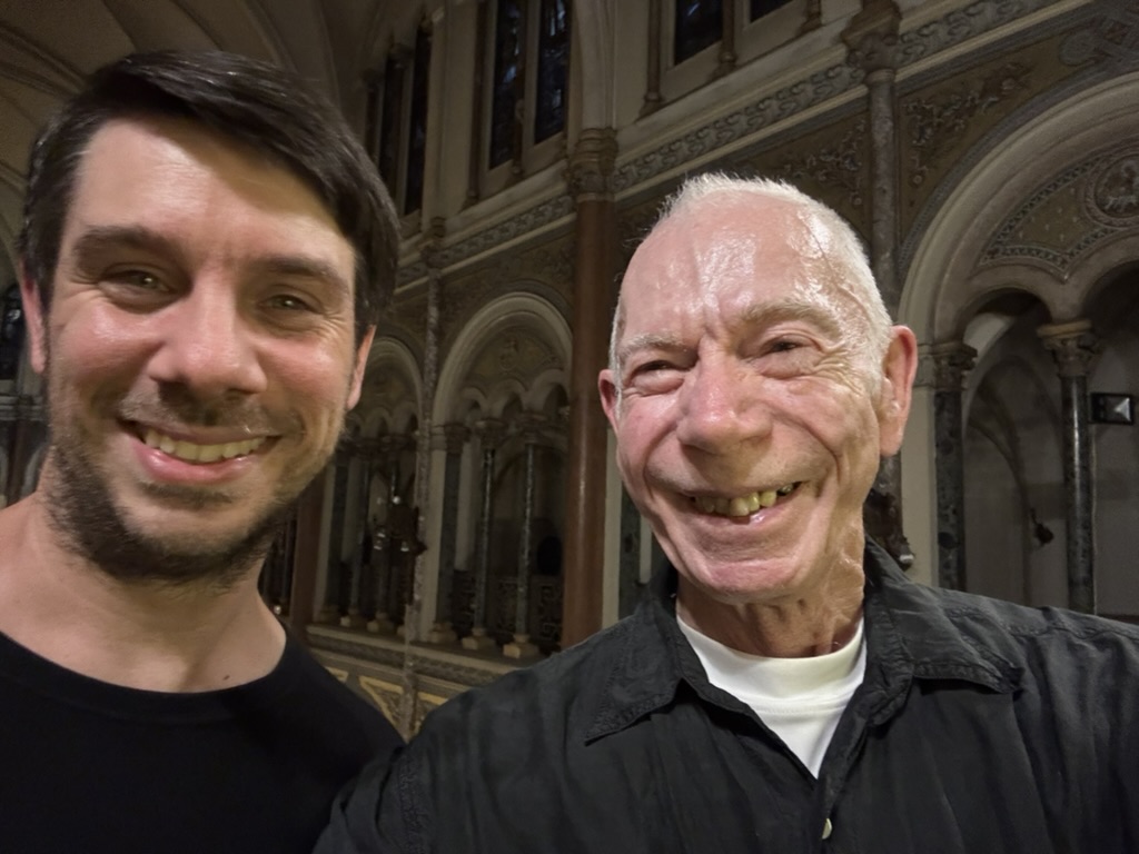 Two men smiling for a selfie in a beautifully ornate indoor setting with architectural details in the background.