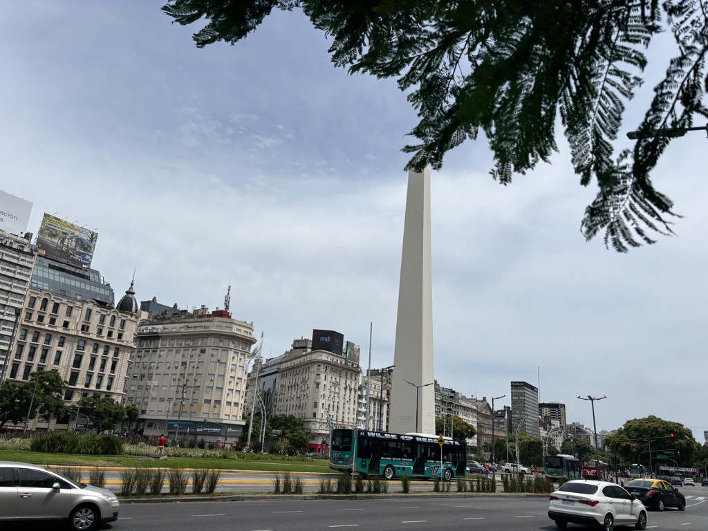 A view of the iconic Obelisk in Buenos Aires, surrounded by historic buildings and city traffic, with green foliage framing the scene.