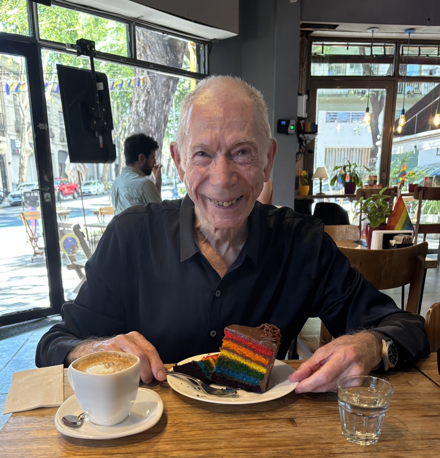 An elderly man with gray hair is smiling while sitting at a café table, holding a plate with a slice of rainbow cake and a fork. A cup of cappuccino and a glass of water are on the table in front of him.