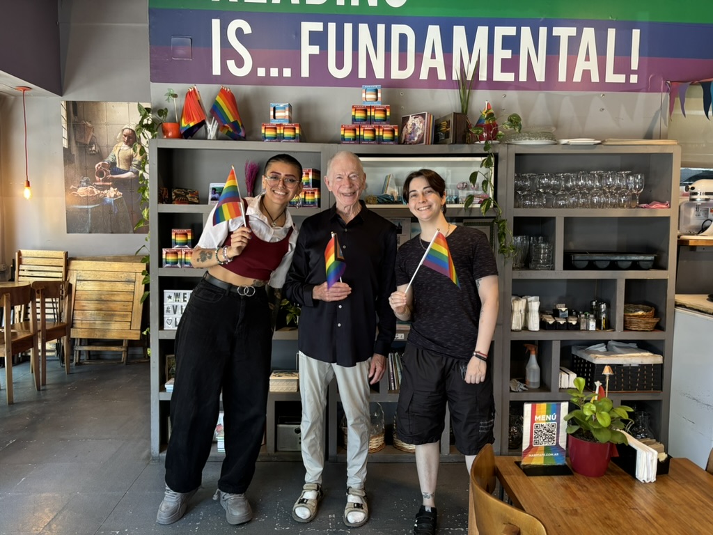Three people standing in a café, all holding small rainbow flags, with a colorful banner reading 'READING IS... FUNDAMENTAL!' in the background.