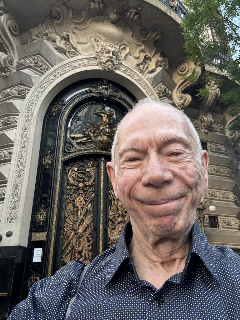 A smiling older man in a patterned shirt stands in front of an ornate decorative building entrance featuring intricate designs and sculptures.