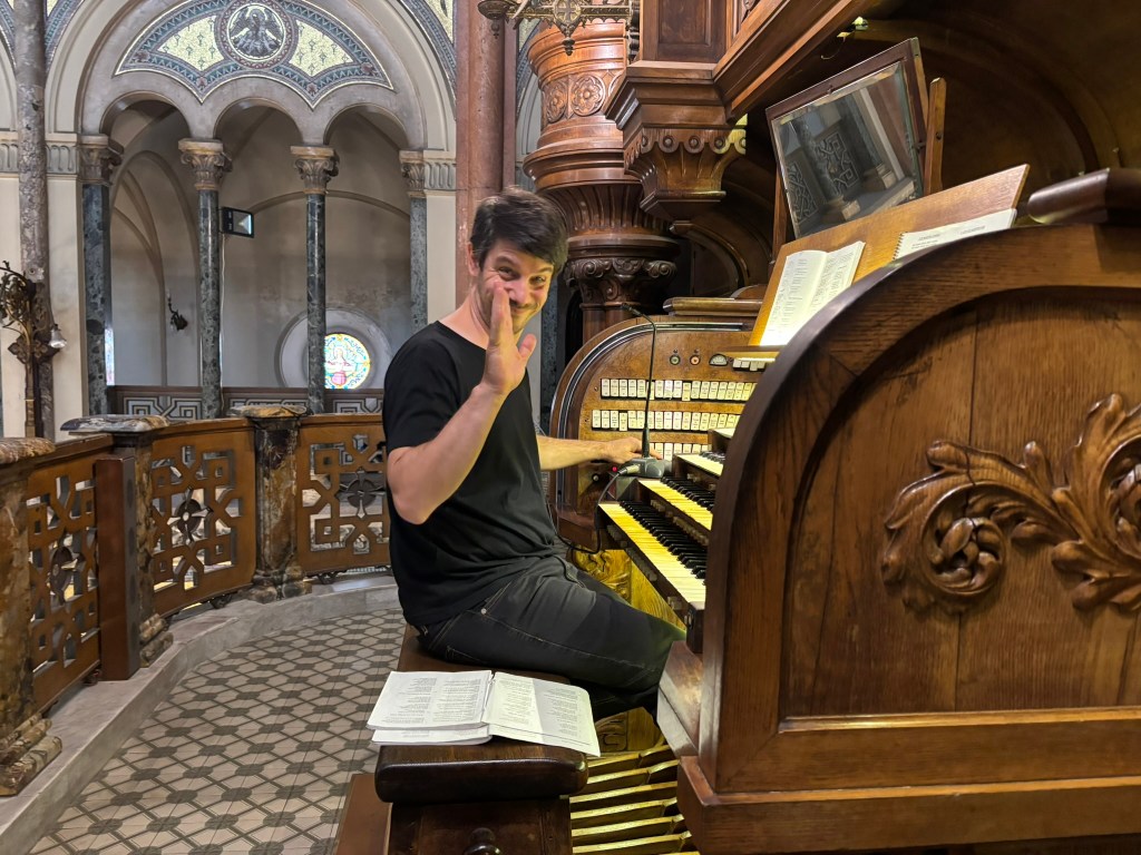 A man playing a large wooden organ inside a church, smiling and waving at the camera, with sheets of music on the organ.