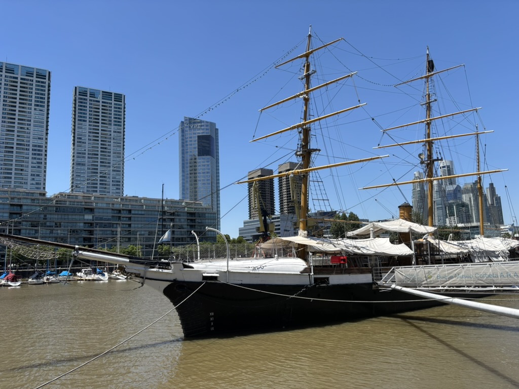 A traditional tall ship docked on a river with modern high-rise buildings in the background.