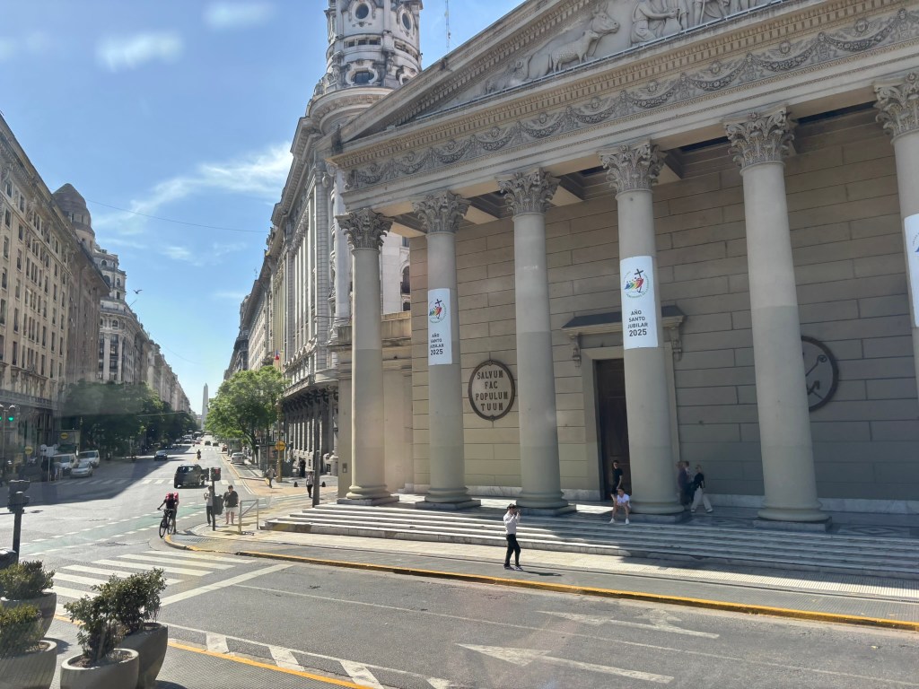 View of a historic building with large columns, featuring banners and sculptures on the facade, situated on a city street with few pedestrians and trees.
