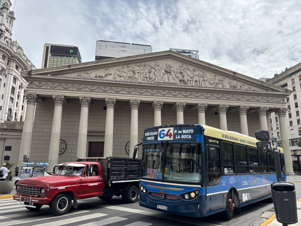 A blue and yellow bus labeled '64' at a busy intersection in front of a classical building with elaborate sculptures and columns, accompanied by a red truck parked nearby.