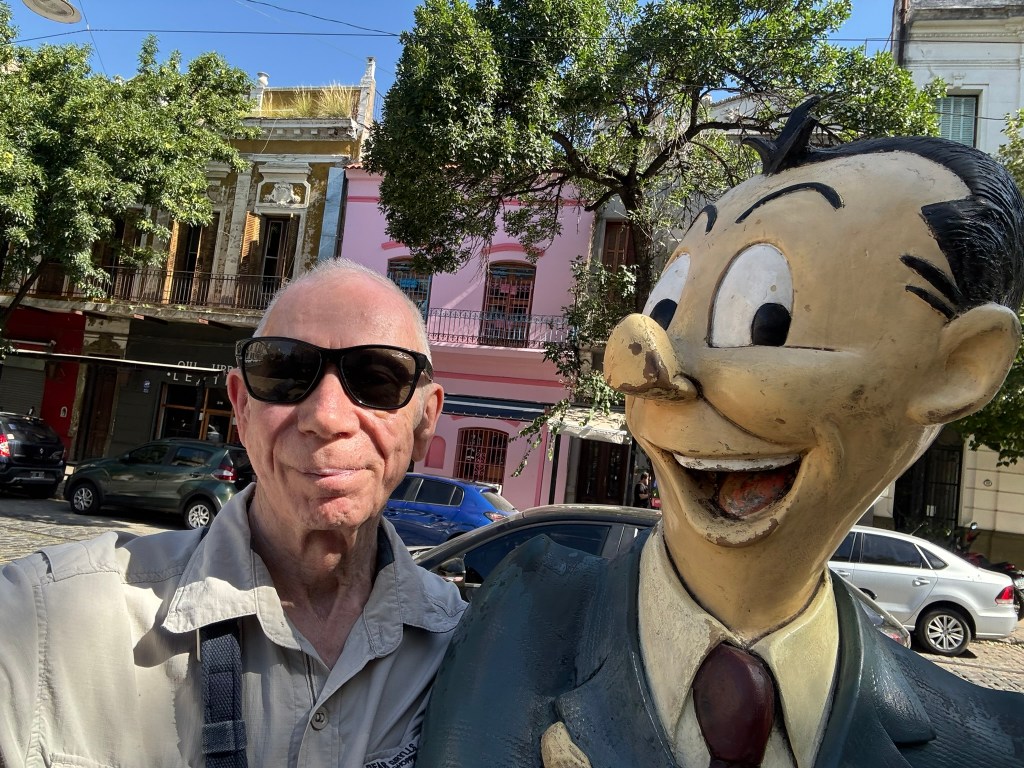 A person wearing sunglasses stands next to a smiling cartoon character statue, with colorful buildings and trees in the background.