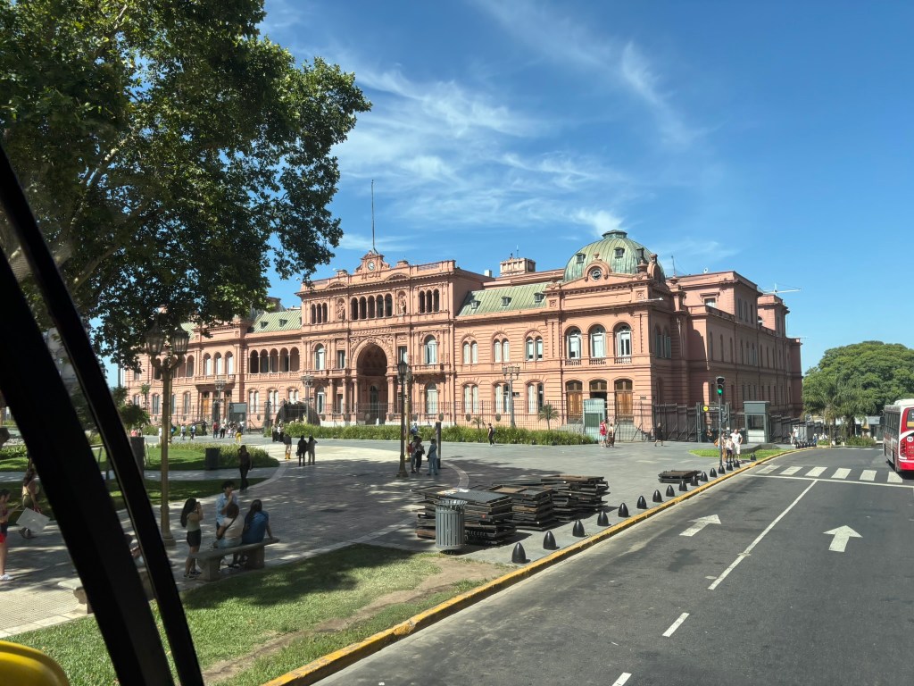 A historical pink building with a dome, surrounded by green trees and people in a plaza.