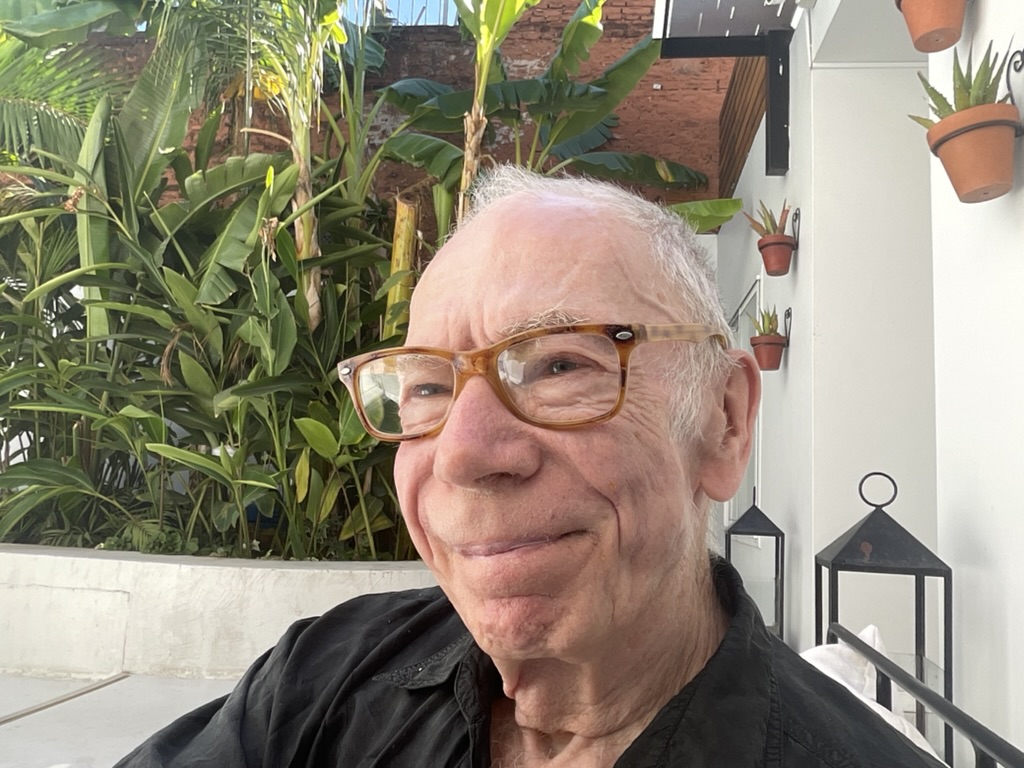 A smiling older man with glasses sitting in front of lush green plants, wearing a black shirt.