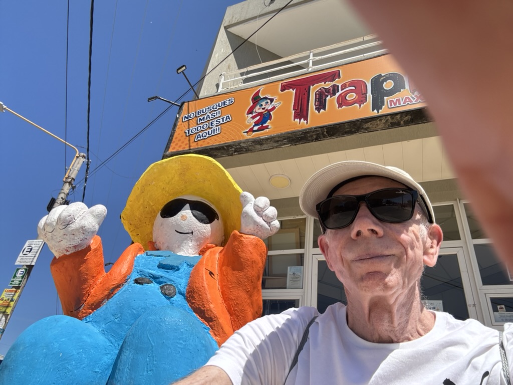 A person taking a selfie with a colorful statue of a cartoon character wearing a large yellow hat and sunglasses, in front of a shop sign.
