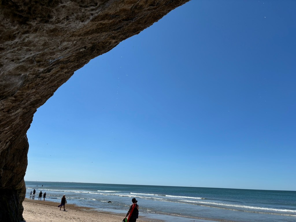 A beach scene viewed from beneath a rocky arch, with clear blue skies and several people walking along the shoreline.