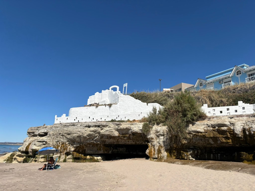 A beach scene featuring a rocky shoreline with a white building on top of the hill against a clear blue sky. A person sitting under a blue and white umbrella is visible on the sandy beach.