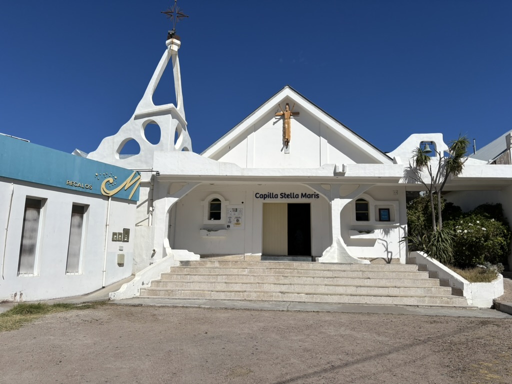 Exterior view of Capilla Stella Maris, a white chapel with a distinctive steeple and a cross, flanked by greenery and clear blue skies.
