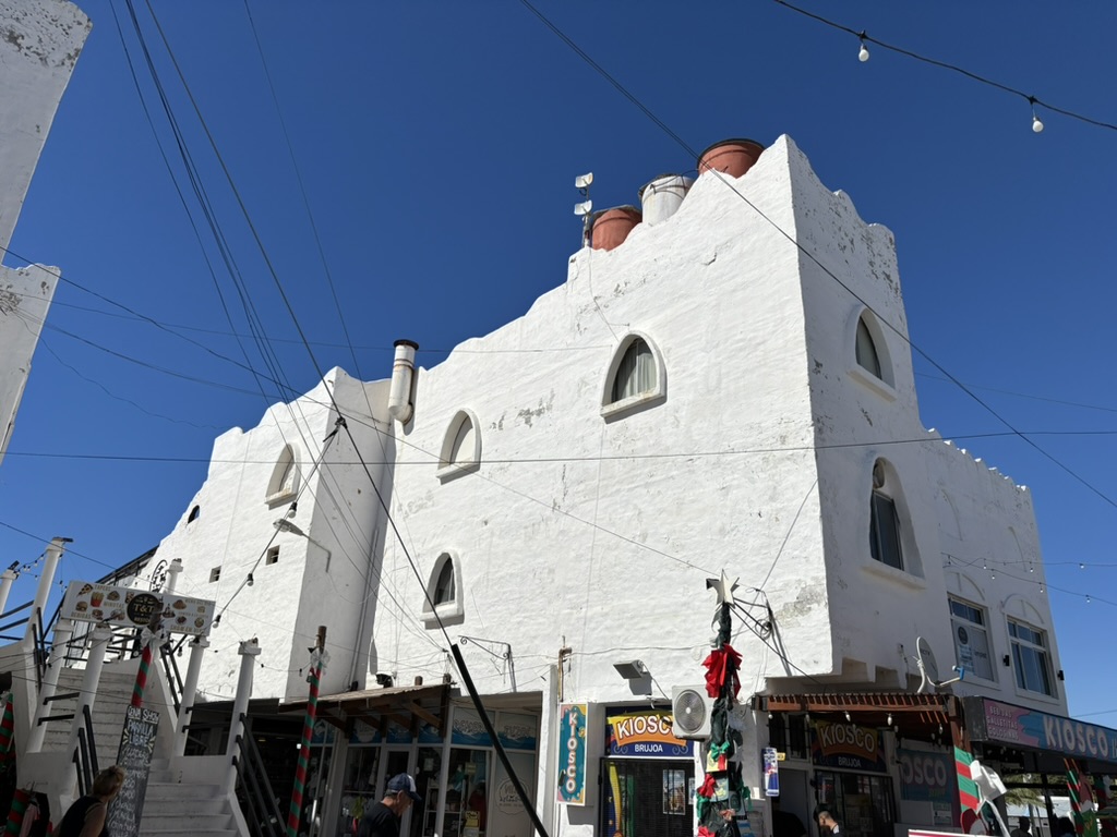 A white building with distinctive arched windows and clay roof tanks, surrounded by shops in a lively outdoor market under a clear blue sky.