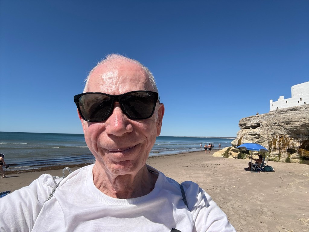 A person wearing sunglasses smiling at the beach, with a clear blue sky and the ocean in the background.