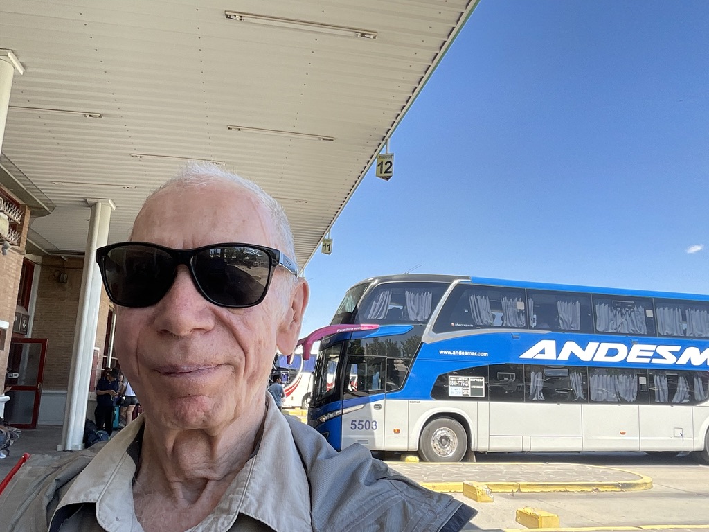 An older man with sunglasses takes a selfie at a bus terminal, with an Andesmar bus parked in the background under a clear blue sky.