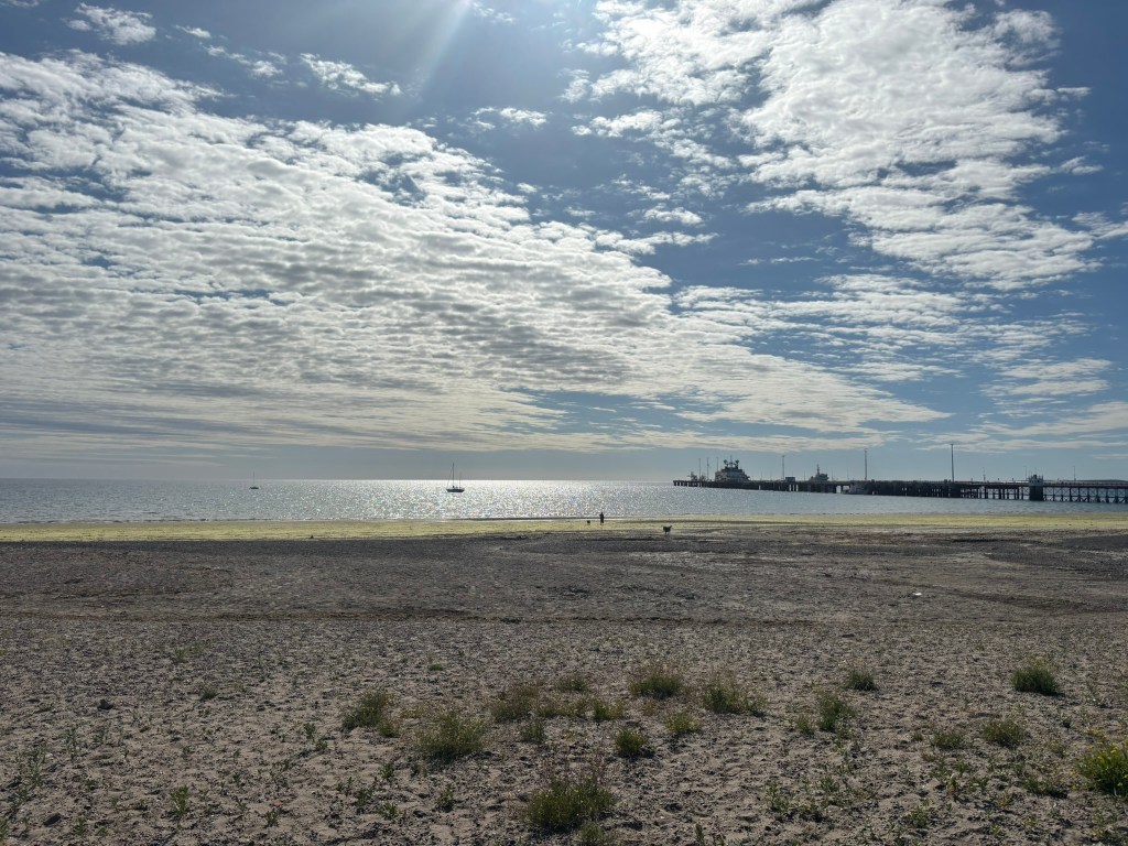 A serene view of a beach with a calm sea reflecting the sunlight, scattered clouds in the sky, and a distant pier with boats anchored near the shore.