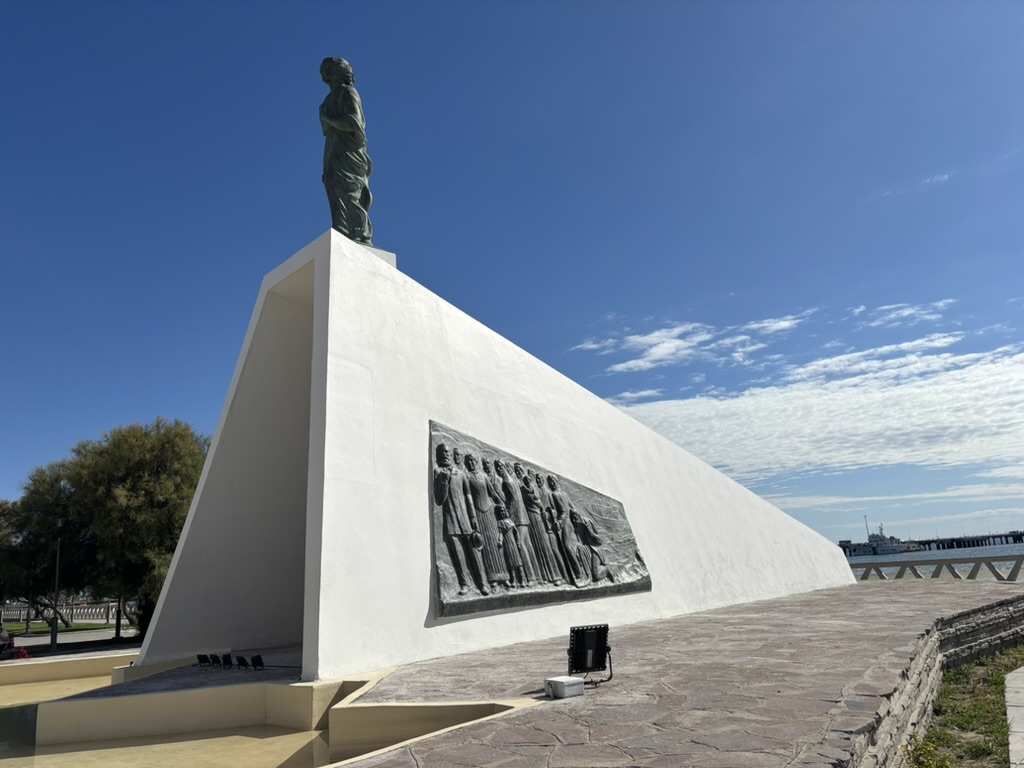 A large white monument featuring a statue on top and a bas-relief depicting figures on one side, set against a clear blue sky.