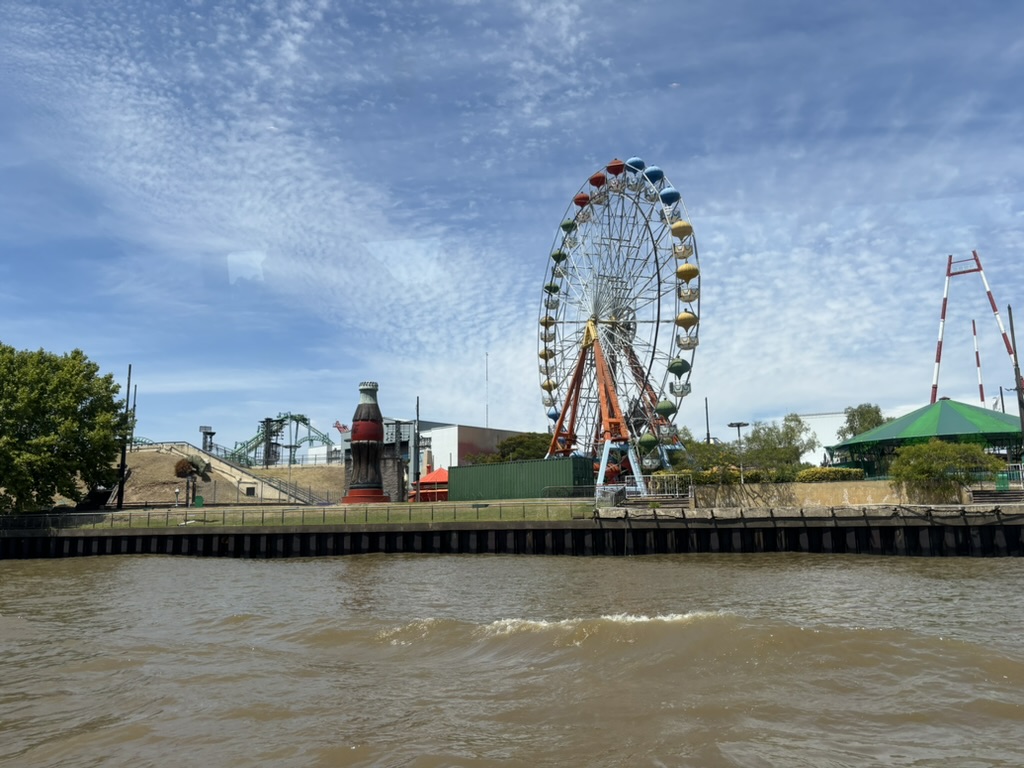 View of a waterfront amusement park featuring a large Ferris wheel, roller coaster tracks, and a giant soft drink bottle structure under a blue sky with clouds.