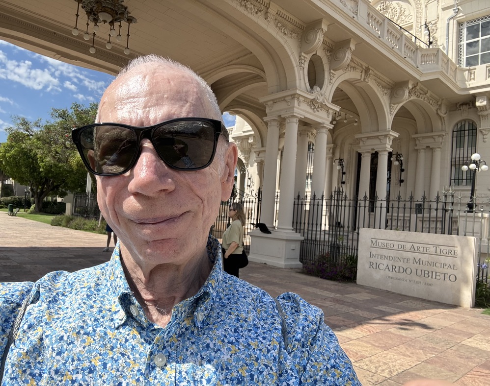 A person with sunglasses smiles for a selfie in front of the Museo de Arte Tigre, showcasing its ornate architecture and a sign with the museum's name.