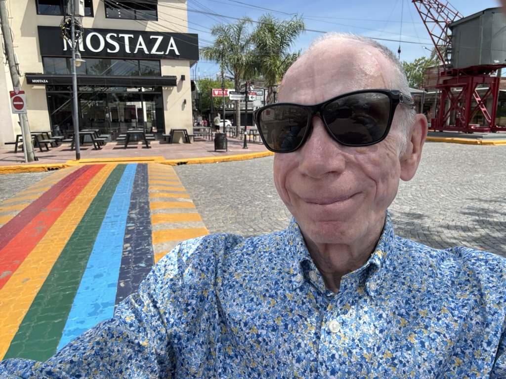 A person wearing sunglasses smiles for a selfie in front of a restaurant named Mostaza, with a colorful rainbow crosswalk in the foreground.