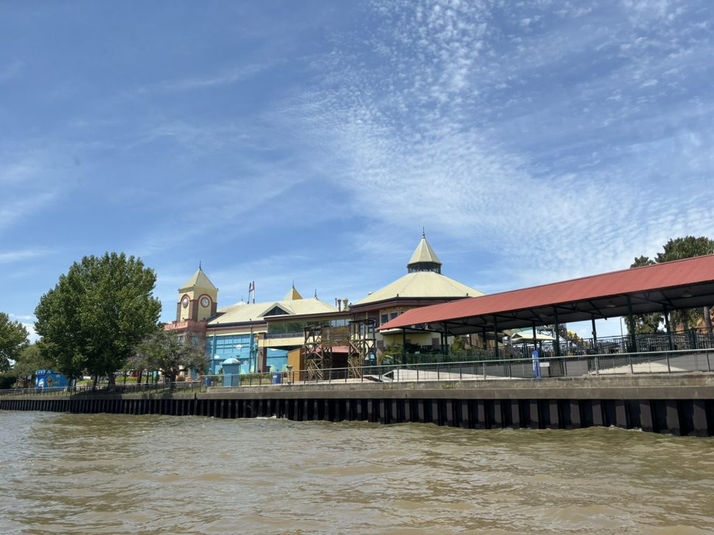 View of a waterfront building with distinct architecture and a clock tower, surrounded by trees under a blue sky.