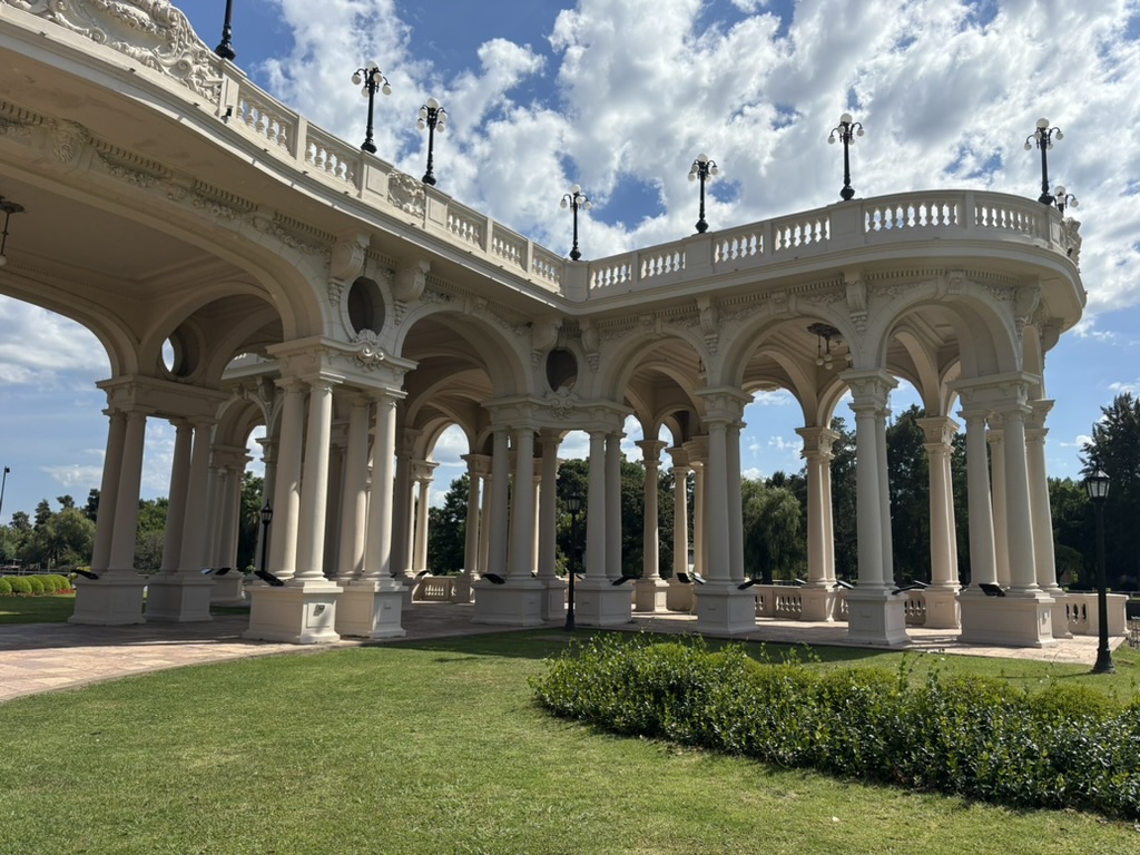 A beautiful architectural structure featuring arched columns, ornate detailing, and decorative lamps, set in a park with green grass and trees under a bright blue sky.