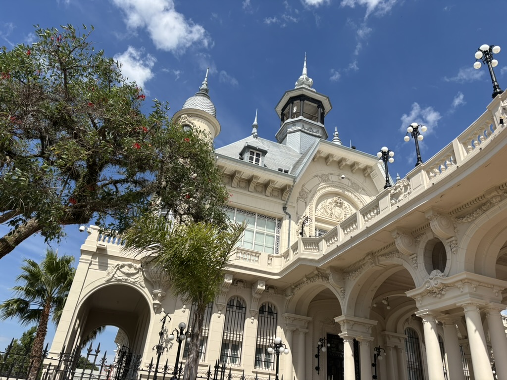 A decorative architectural building with a grey roof and spire, featuring intricate details and arched columns, surrounded by trees and blue sky.