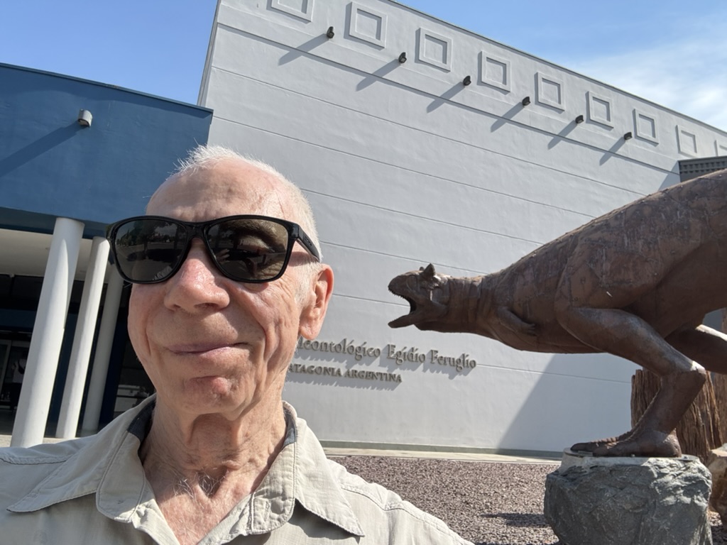 A man wearing sunglasses poses in front of a paleontological museum with a dinosaur statue.